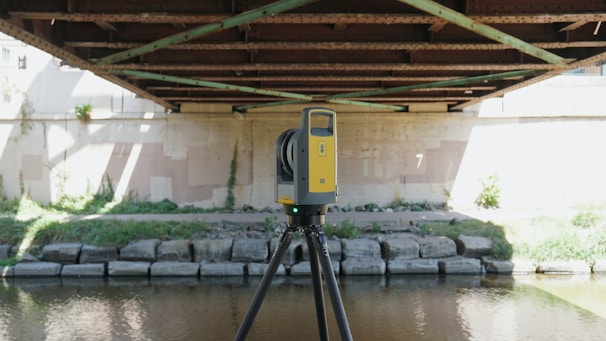 A yellow and black tripod-mounted surveying instrument is positioned under a metal bridge by a riverbank. The bridge has rust-colored girders and shadows cast across a concrete wall. Lush greenery grows along the banks with patches of sunlight highlighting parts of the scene.