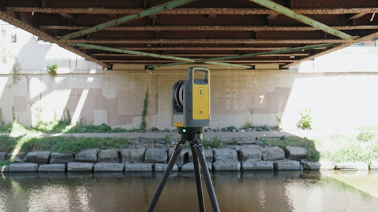 A yellow and black tripod-mounted surveying instrument is positioned under a metal bridge by a riverbank. The bridge has rust-colored girders and shadows cast across a concrete wall. Lush greenery grows along the banks with patches of sunlight highlighting parts of the scene.
