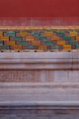 A section of a decorative wall featuring a mix of geometric and ornate patterns. The upper part consists of colorful rectangular tiles in green, yellow, and brown, arranged in a brick-like pattern. Below, there is an ornately carved stone or marble surface with intricate scroll designs.
