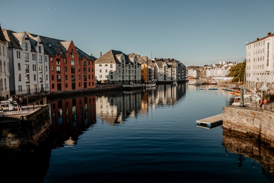 A canal lined with colorful, historic buildings reflecting in the calm water. The scene is set in a picturesque town with clear skies and gentle lighting, highlighting the distinct architecture. A few people are visible near the waterfront, enjoying the serene atmosphere.