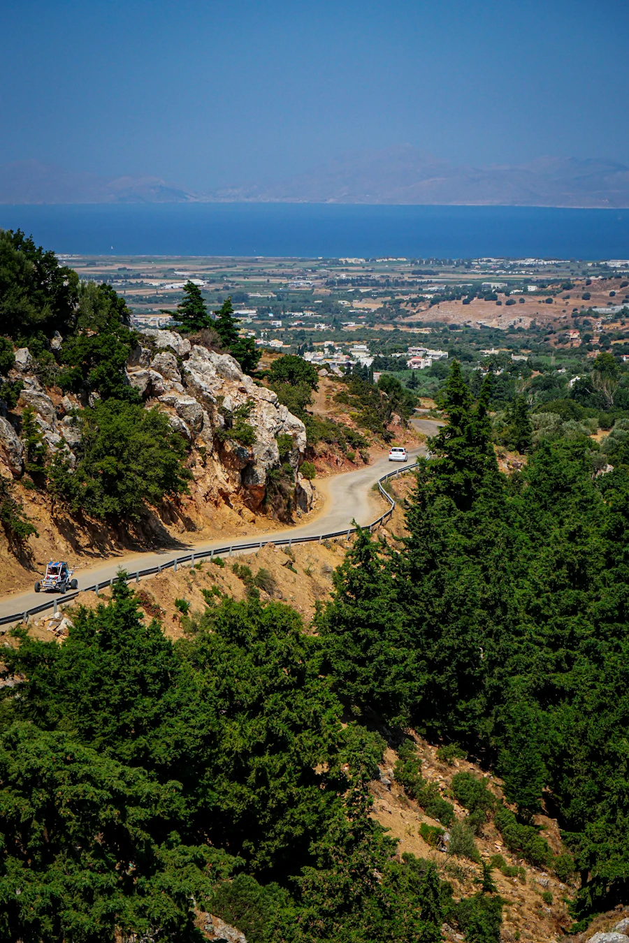 A coast road climbing across a steep green hillside above the sea, with a small open vehicle on the lower bend and hazy islands across the water.