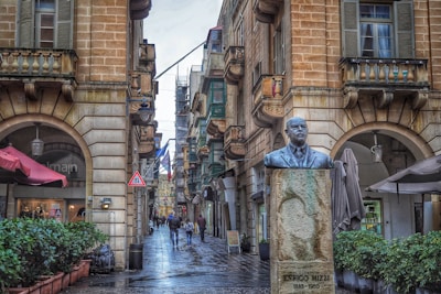 A narrow cobblestone street lined with historic buildings featuring balconies and arches. A bust of Enrico Mizzi stands prominently on a pedestal in the foreground. Pedestrians walk along the street, some carrying bags. The scene includes a variety of flags and signs, indicating a lively urban environment. Potted plants and patio umbrellas add a touch of greenery.