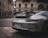 A sleek silver sports car is parked on a European city street. The architecture of the surrounding buildings features classic stone facades with arched windows. A person walks in the background, and the street is lined with cobblestones.