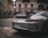 A sleek silver sports car is parked on a European city street. The architecture of the surrounding buildings features classic stone facades with arched windows. A person walks in the background, and the street is lined with cobblestones.