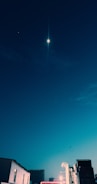 A night sky featuring a bright celestial object and a faint star surrounded by deep blue hues. Below, urban buildings are partially illuminated, hinting at a serene and quiet cityscape.