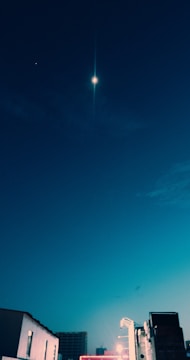 A night sky featuring a bright celestial object and a faint star surrounded by deep blue hues. Below, urban buildings are partially illuminated, hinting at a serene and quiet cityscape.