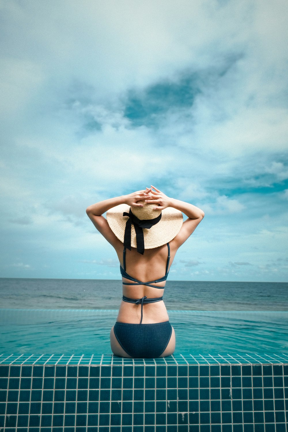 a woman in a bikini and hat sitting on the edge of a swimming pool