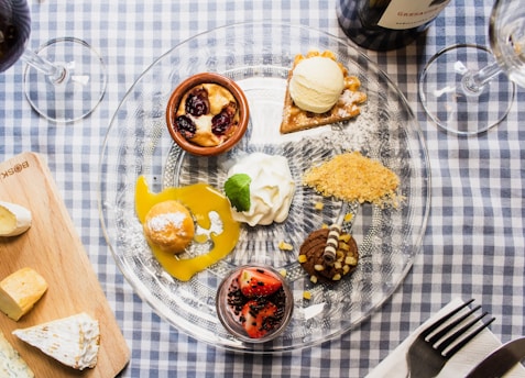 An elegant dessert platter featuring a variety of sweet treats arranged on a clear glass plate. The plate sits on a blue and white checkered tablecloth and includes items such as a small tart with vanilla ice cream, a chocolate dessert with a decorative chocolate curl, a mini torte topped with a cherry, and a dollop of whipped cream garnished with a mint leaf. Adjacent to the plate are a wooden board with assorted cheeses and glasses of red wine, creating an inviting and luxurious setting.