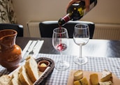 A table is set with a selection of cheeses on a wooden board and slices of bread in a basket. A person is pouring red wine into a wine glass. There are two empty wine glasses and a brown ceramic jug nearby. Silverware is neatly arranged on a checked tablecloth.