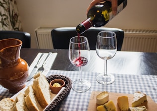 A table is set with a selection of cheeses on a wooden board and slices of bread in a basket. A person is pouring red wine into a wine glass. There are two empty wine glasses and a brown ceramic jug nearby. Silverware is neatly arranged on a checked tablecloth.