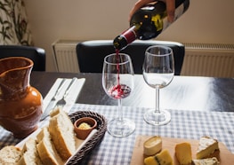 A table is set with a selection of cheeses on a wooden board and slices of bread in a basket. A person is pouring red wine into a wine glass. There are two empty wine glasses and a brown ceramic jug nearby. Silverware is neatly arranged on a checked tablecloth.