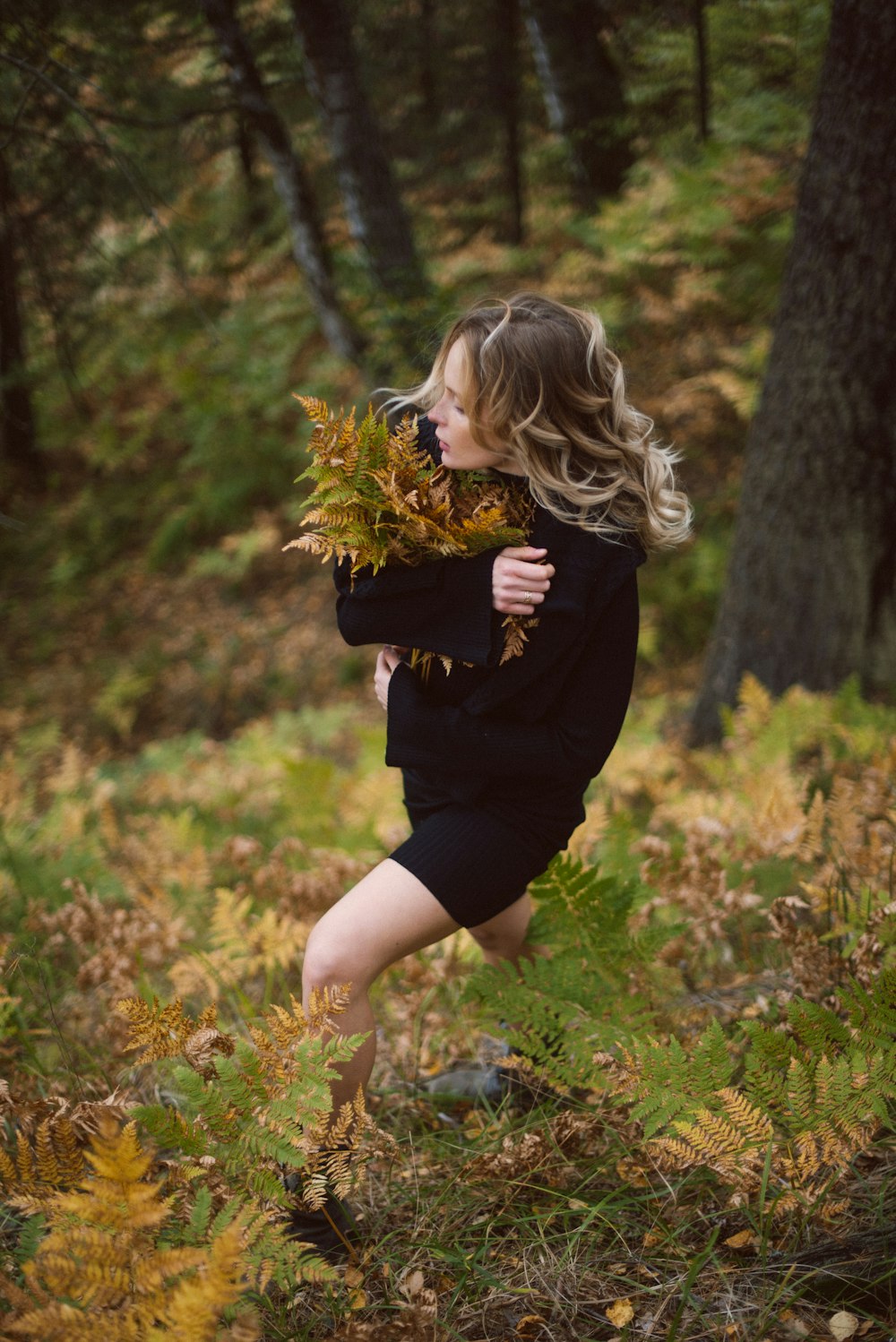 woman wearing black crew-neck long-sleeved dress holding green and brown fern plant while walking near trees during daytime