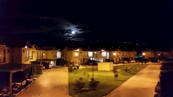 A residential neighborhood at night with a clear view of the moon partially obscured by clouds. Streetlights illuminate the area, casting a warm glow over the parked cars and well-maintained lawns. Rows of similar looking houses line both sides of the street.