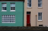 A street view of two adjacent houses with contrasting colors. The house on the left is painted in a vibrant turquoise, featuring three white-framed windows, one large window with curtains, and two smaller windows above. The house on the right has a neutral beige color with a striking red door at the center and two white-framed windows. A small signpost is positioned near the red door.