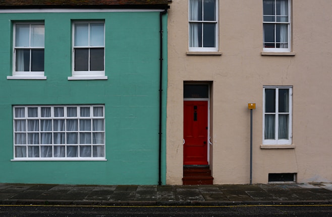 A street view of two adjacent houses with contrasting colors. The house on the left is painted in a vibrant turquoise, featuring three white-framed windows, one large window with curtains, and two smaller windows above. The house on the right has a neutral beige color with a striking red door at the center and two white-framed windows. A small signpost is positioned near the red door.