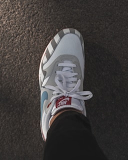 A stylish Nike sneaker with a blend of white, grey, and blue colors is positioned on a textured surface. The sneaker features a red tag on the tongue, matching laces, and is worn with dark pants.