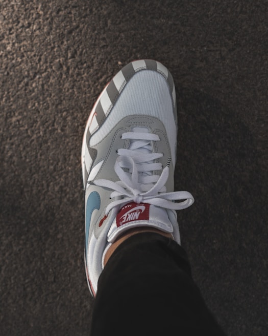 A stylish Nike sneaker with a blend of white, grey, and blue colors is positioned on a textured surface. The sneaker features a red tag on the tongue, matching laces, and is worn with dark pants.
