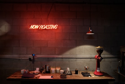A dimly lit coffee shop setting featuring a wooden table against a concrete wall with a neon sign reading 'NOW ROASTING.' The table holds a variety of coffee equipment including cups, a tray, a kettle, and a coffee grinder. A single overhead light casts a warm glow over the scene, enhancing the inviting atmosphere.