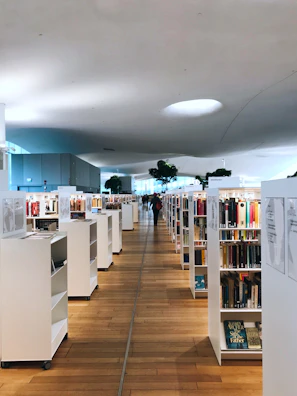 A spacious, modern library with wooden flooring and white bookshelves filled with books. The ceiling has an organic shape with soft lighting. A person is walking in the distance, adding depth to the scene. Green potted plants are visible among the shelves, contributing to a serene atmosphere.