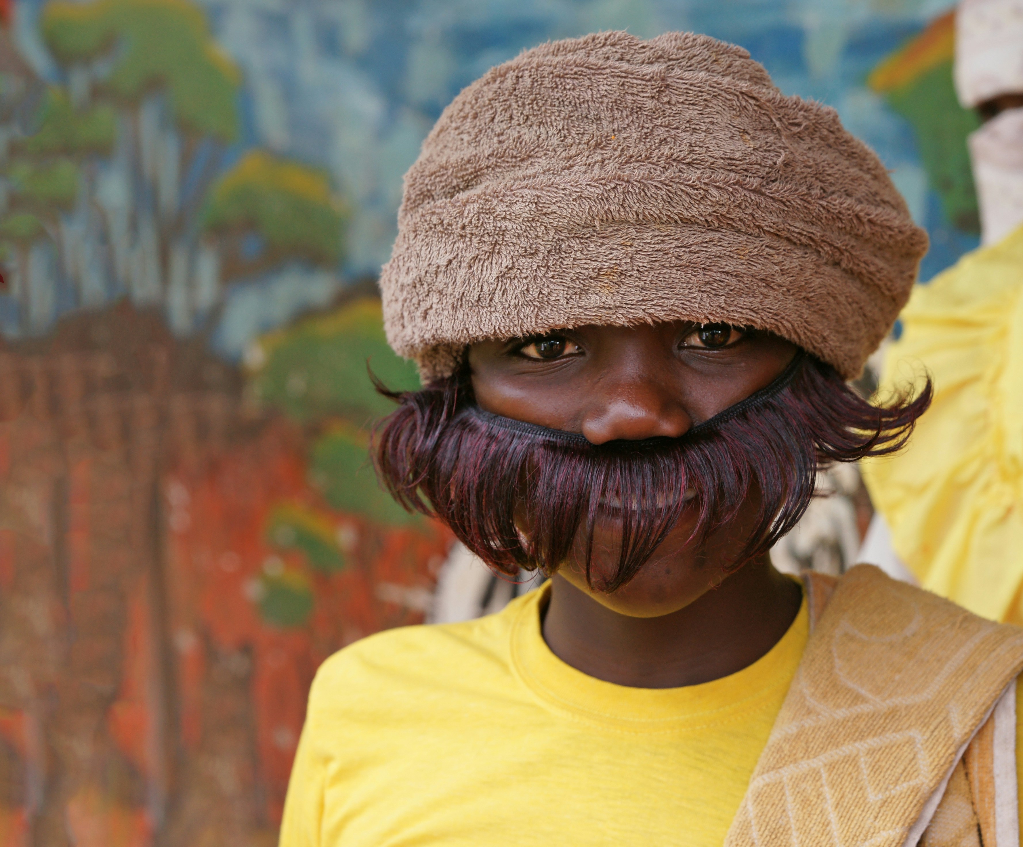 Portrait of a person wearing a fuzzy brown hat and yellow shirt, set against a vivid painted backdrop. The eyes anchor the frame, and a playful hat adds character.