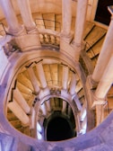 A spiral staircase with multiple levels, viewed from above, with columns evenly spaced around the edges. The balustrades and railing add to the architectural elegance, and light reflects off the stone surfaces, creating a warm ambiance.