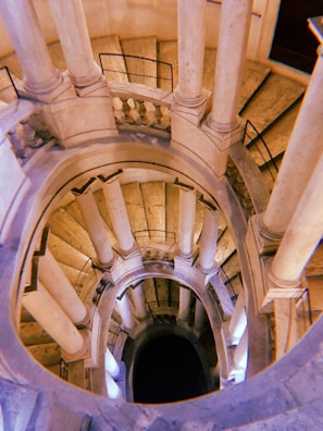 A spiral staircase with multiple levels, viewed from above, with columns evenly spaced around the edges. The balustrades and railing add to the architectural elegance, and light reflects off the stone surfaces, creating a warm ambiance.