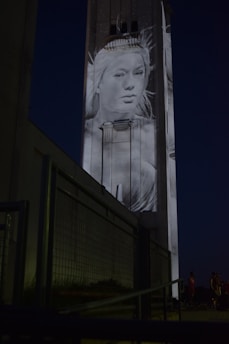 A large building at night with a monochromatic projection of a woman's face and shoulders. The image is displayed on the tall structure, making it prominent against the dark sky. In the foreground, there is a metal railing leading towards the building.