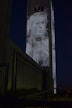 A large building at night with a monochromatic projection of a woman's face and shoulders. The image is displayed on the tall structure, making it prominent against the dark sky. In the foreground, there is a metal railing leading towards the building.