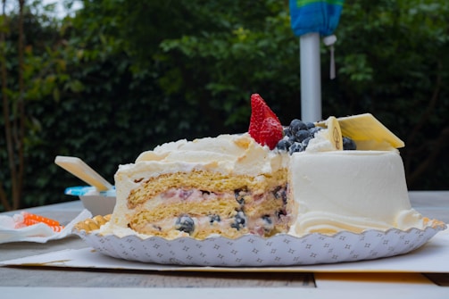 A partially sliced cake with multiple layers of cream and sponge, topped with fresh blueberries and a single strawberry. The cake is placed on a decorative paper plate. In the background, there is greenery suggesting an outdoor setting, along with a folded umbrella.