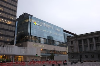A modern urban street scene featuring a glass office building with the Microsoft logo and a store labeled Nordstrom. The area includes an outdoor seating arrangement with red chairs and tables. The weather appears overcast, creating a subdued atmosphere. Adjacent buildings exhibit contemporary and classical architectural styles.