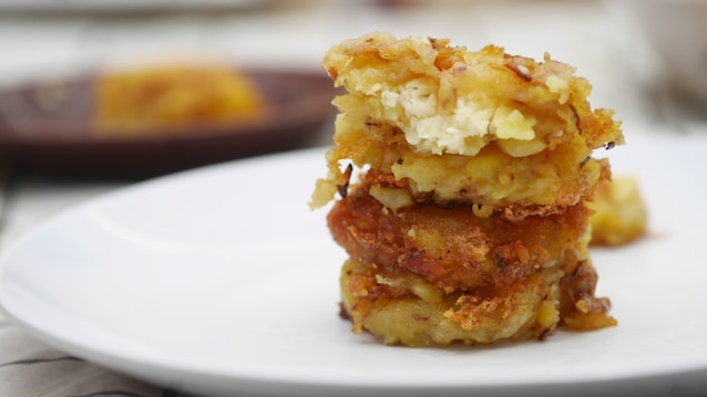 A stack of three crispy, golden-brown fritters is placed on a white plate. The fritters appear to have a creamy filling, possibly cheese, visible through the top layer. The background is softly blurred, with a hint of another dish in the distance, creating a focus on the fritters.