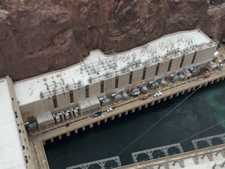 A large industrial structure is positioned against a rocky cliffside next to a body of water. The building is equipped with numerous electrical transformers and cables on its roof. The architecture features a long, rectangular shape with multiple vertical windows. A walkway with vehicles and equipment is visible next to the water.