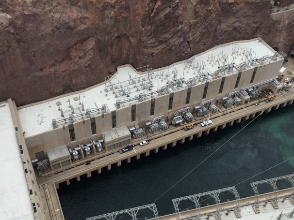 A large industrial structure is positioned against a rocky cliffside next to a body of water. The building is equipped with numerous electrical transformers and cables on its roof. The architecture features a long, rectangular shape with multiple vertical windows. A walkway with vehicles and equipment is visible next to the water.