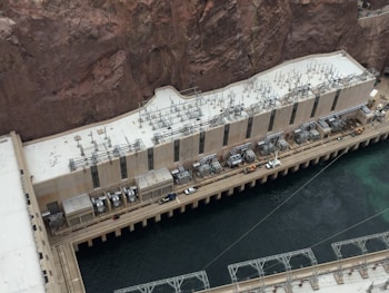 A large industrial structure is positioned against a rocky cliffside next to a body of water. The building is equipped with numerous electrical transformers and cables on its roof. The architecture features a long, rectangular shape with multiple vertical windows. A walkway with vehicles and equipment is visible next to the water.