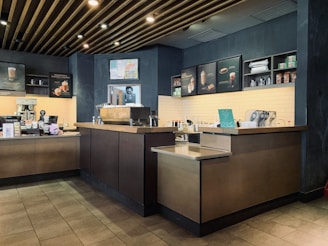 A modern coffee shop interior featuring a coffee preparation counter with various equipment, including a coffee machine, cups, and containers. The wall has a menu display with images of drinks and snacks. The overall decor includes dark wooden accents and a tiled floor.