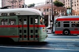 Two public transit vehicles are parked on a city street surrounded by tall buildings. The vehicle on the left is a vintage-style green and red streetcar, while the one on the right is a modern gray and red city bus. Overhead wires and street signs are visible, along with an ornate building facade and trees in the background.