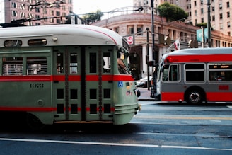 Two public transit vehicles are parked on a city street surrounded by tall buildings. The vehicle on the left is a vintage-style green and red streetcar, while the one on the right is a modern gray and red city bus. Overhead wires and street signs are visible, along with an ornate building facade and trees in the background.