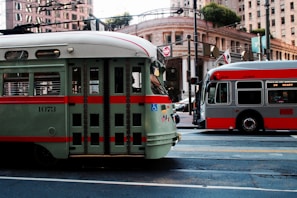 Two public transit vehicles are parked on a city street surrounded by tall buildings. The vehicle on the left is a vintage-style green and red streetcar, while the one on the right is a modern gray and red city bus. Overhead wires and street signs are visible, along with an ornate building facade and trees in the background.