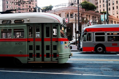 Two public transit vehicles are parked on a city street surrounded by tall buildings. The vehicle on the left is a vintage-style green and red streetcar, while the one on the right is a modern gray and red city bus. Overhead wires and street signs are visible, along with an ornate building facade and trees in the background.