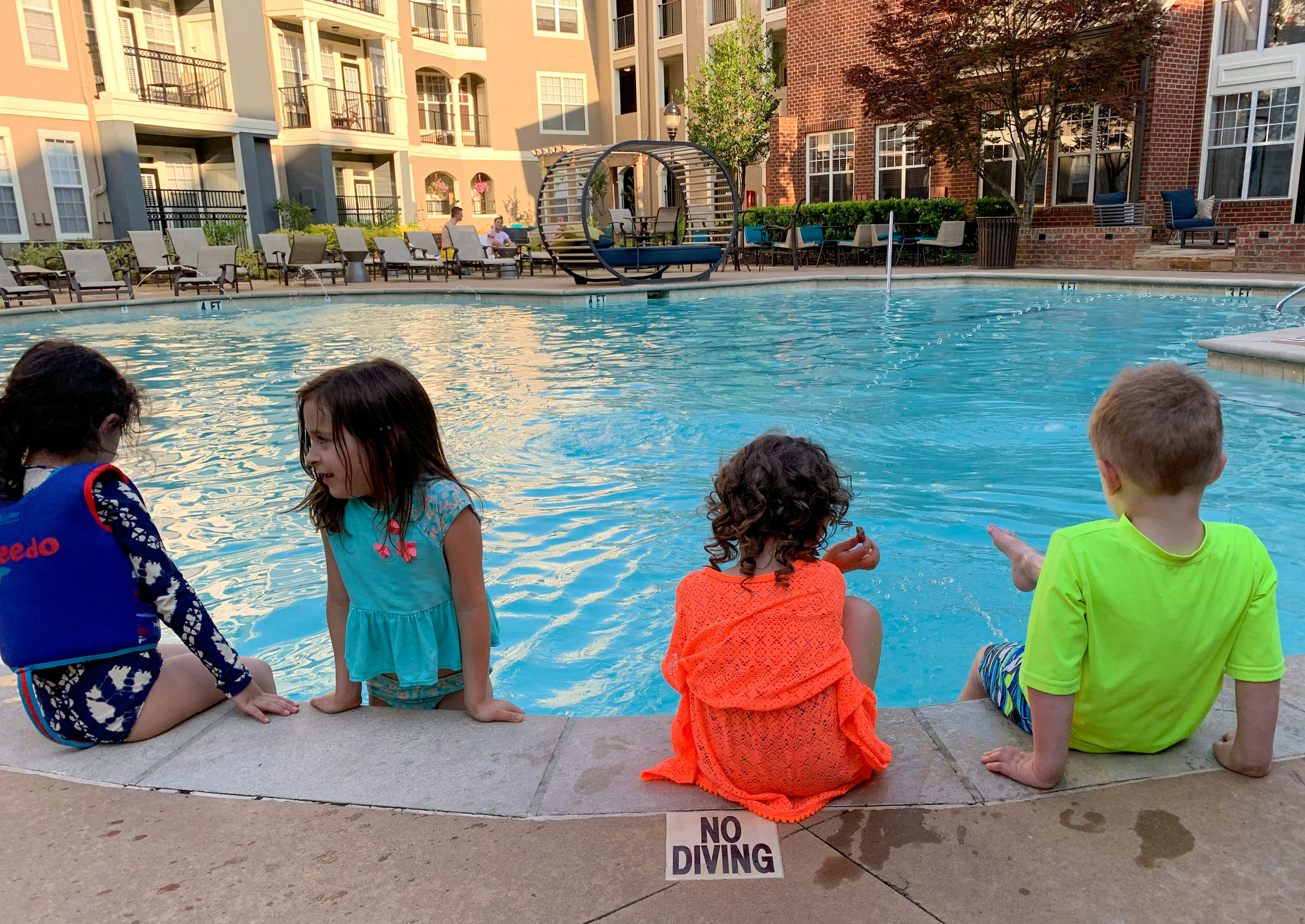 Four children sit by the edge of a swimming pool, with their feet in the water. They are surrounded by apartment buildings and poolside furniture including chairs and loungers. The setting is a communal outdoor area with a clear and calm pool. A 'No Diving' sign is visible on the pool's edge.