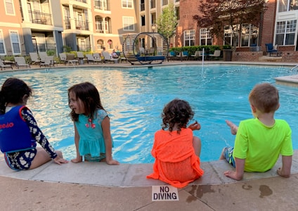 Four children sit by the edge of a swimming pool, with their feet in the water. They are surrounded by apartment buildings and poolside furniture including chairs and loungers. The setting is a communal outdoor area with a clear and calm pool. A 'No Diving' sign is visible on the pool's edge.