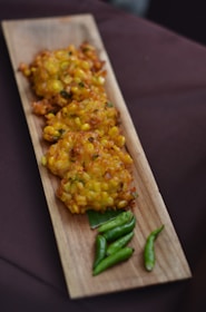 Crispy corn fritters are arranged on a rectangular wooden serving tray, accompanied by a few vibrant green chilies. The fritters appear golden brown with visible corn kernels, giving a rustic and appetizing appearance.