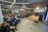 A large group of people are sitting in a modern, well-lit room, attentively watching a panel discussion. The room features contemporary design elements, including bright overhead lighting and wood panel accents on the walls. Several people are seated on chairs, while a few are on a raised platform as speakers.