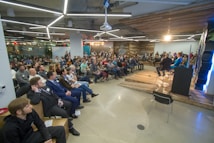 A large group of people are sitting in a modern, well-lit room, attentively watching a panel discussion. The room features contemporary design elements, including bright overhead lighting and wood panel accents on the walls. Several people are seated on chairs, while a few are on a raised platform as speakers.