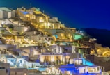 A picturesque hillside with whitewashed buildings illuminated at night, displaying classical Greek architecture. The structures are stacked in tiers, creating a captivating aesthetic against a deep blue sky. Soft, warm lighting from windows and outdoor fixtures adds a cozy and inviting feel.