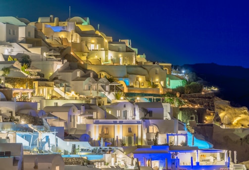 A picturesque hillside with whitewashed buildings illuminated at night, displaying classical Greek architecture. The structures are stacked in tiers, creating a captivating aesthetic against a deep blue sky. Soft, warm lighting from windows and outdoor fixtures adds a cozy and inviting feel.