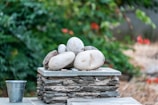A small stack of smooth, rounded stones rests on a flat, stone base, surrounded by a lush background of green foliage. To the side, a metal bucket sits on a surface made of similar stone material, adding a rustic touch to the serene setup. The focus is on the stones which are varying in size and color, creating a natural, balanced arrangement.