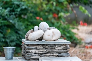 A small stack of smooth, rounded stones rests on a flat, stone base, surrounded by a lush background of green foliage. To the side, a metal bucket sits on a surface made of similar stone material, adding a rustic touch to the serene setup. The focus is on the stones which are varying in size and color, creating a natural, balanced arrangement.