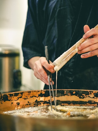 A person wearing dark clothing holds a pair of metal tongs and a long stick, dipping food into a large, bubbling copper pot filled with oil. The setting suggests a kitchen or cooking environment, focusing on a culinary process.