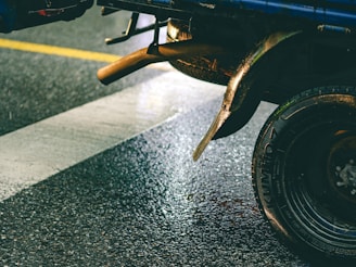 A close-up shot of the rear wheel and exhaust pipe of a vehicle parked on a wet road. The vehicle is positioned on a zebra crossing, and the road surface appears shiny due to rainfall. The tire is labeled with a visible brand name, and the metallic parts show some signs of rust.